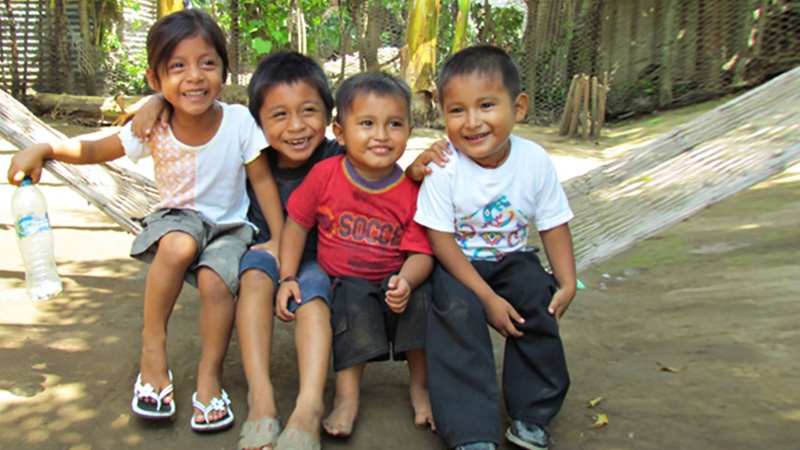 A group of four kids sitting on a hammock over a dirt patch.
